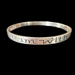 May include: A silver-colored bangle bracelet with the words "I am with" engraved around the outside. The bracelet has a simple, minimalist design and is set against a black background.