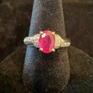May include: A silver-toned ring featuring a large, oval-cut, vibrant pink gemstone at the center. The band is accented with small, clear, round-cut stones embedded along the shoulders on both sides of the central setting, held by four prongs.