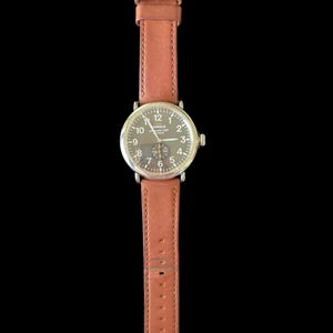 K&ouml;nnte beinhalten: Eine Armbanduhr mit braunem Lederarmband und silberfarbenem Geh&auml;use. Das Zifferblatt der Uhr ist dunkel mit wei&szlig;en Ziffern und Zeigern. Das Armband wird mit einer silbernen Schnalle befestigt.