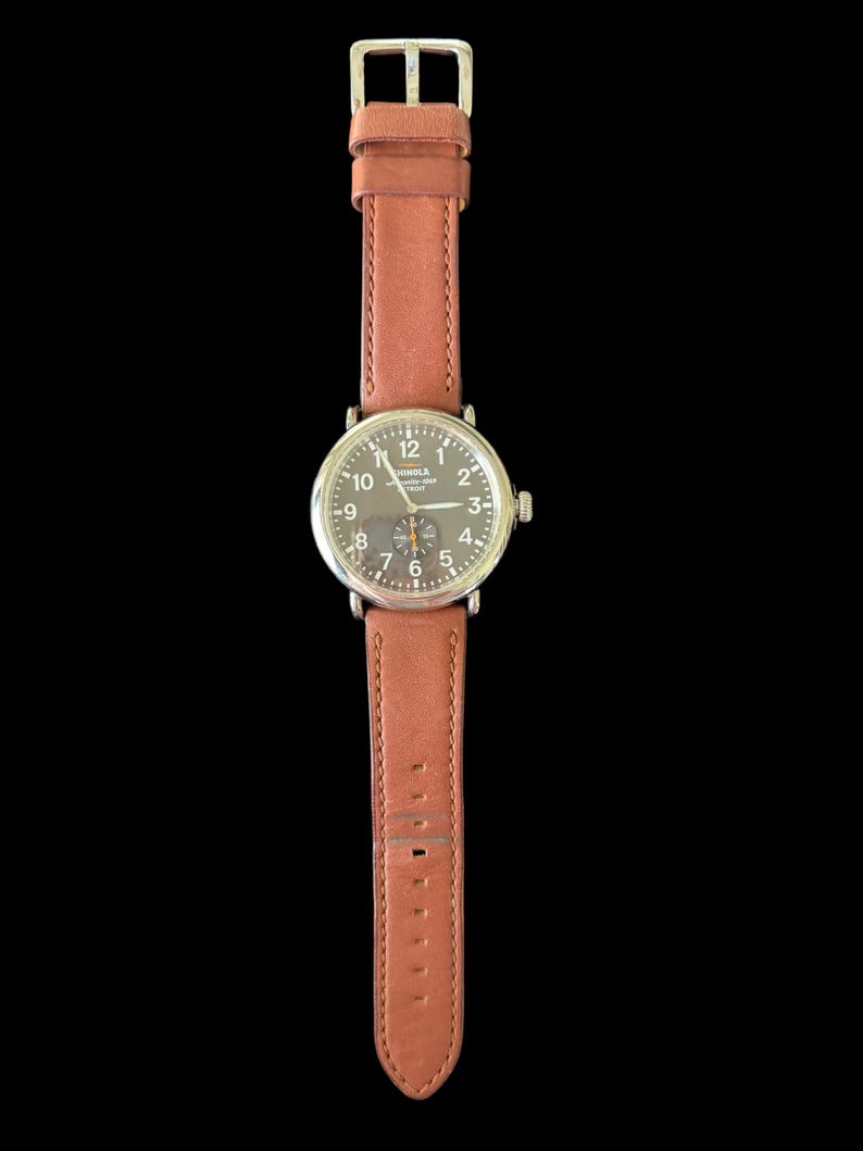 K&ouml;nnte beinhalten: Eine Armbanduhr mit braunem Lederarmband und silberfarbenem Geh&auml;use. Das Zifferblatt der Uhr ist dunkel mit wei&szlig;en Ziffern und Zeigern. Das Armband wird mit einer silbernen Schnalle befestigt.