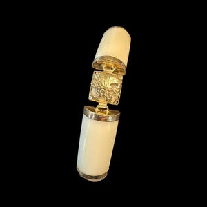 May include: Ivory-colored cylindrical pendant with gold-colored accents. The pendant is composed of two ivory-colored cylinders connected by a gold-colored square with an ornate design. The pendant is set against a black background.