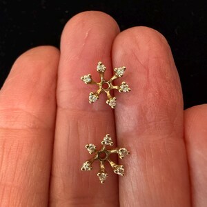 May include: A pair of gold-colored snowflake earrings, each with six small, clear gemstones. The earrings are displayed against a neutral background.