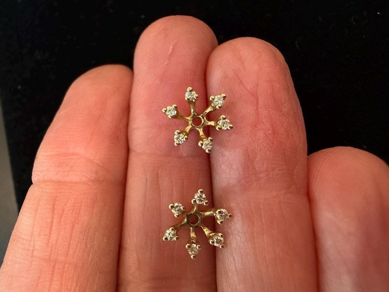 May include: A pair of gold-colored snowflake earrings, each with six small, clear gemstones. The earrings are displayed against a neutral background.
