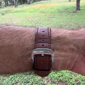 K&ouml;nnte beinhalten: Ein braunes Leder-Uhrenarmband mit silberner Schnalle ist am Handgelenk befestigt. Das Armband ist gen&auml;ht und hat mehrere L&ouml;cher zur Anpassung. Im Hintergrund sind eine gr&uuml;ne Wiese und B&auml;ume zu sehen.