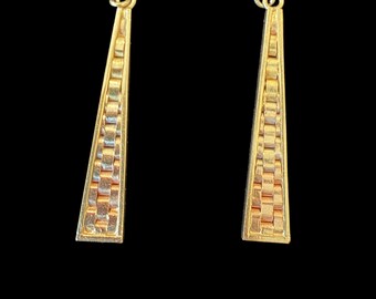 May include: A pair of gold-colored earrings with a geometric, textured design. The earrings are elongated, triangular in shape, and feature a woven pattern. The background is solid black.