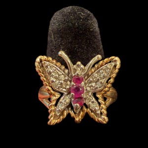 May include: A gold and silver butterfly ring with clear and red gemstones. The ring features a detailed butterfly design with a twisted rope-like border. The butterfly's wings are adorned with clear gemstones, and the body has three red gemstones.