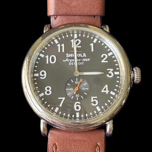 K&ouml;nnte beinhalten: Eine runde Armbanduhr mit braunem Lederarmband. Das Zifferblatt ist dunkelgrau mit wei&szlig;en Ziffern und Zeigern. Auf dem Zifferblatt steht der Text "SHI OLA Argo te-1069 DET OIT".