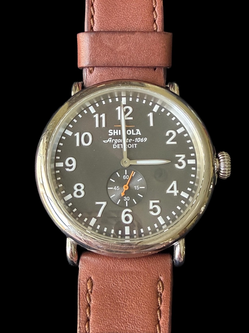 K&ouml;nnte beinhalten: Eine runde Armbanduhr mit braunem Lederarmband. Das Zifferblatt ist dunkelgrau mit wei&szlig;en Ziffern und Zeigern. Auf dem Zifferblatt steht der Text "SHI OLA Argo te-1069 DET OIT".