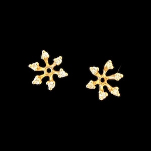 May include: A pair of gold-colored snowflake stud earrings. Each earring features six small, clear gemstones set around a central circular design. The earrings are displayed against a black background.