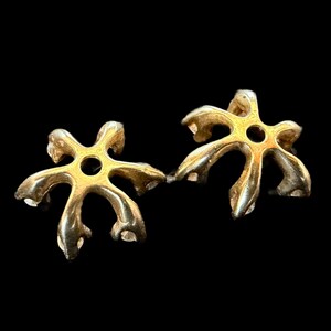 May include: Two gold-colored, star-shaped jewelry components with a central hole. The components have a unique, organic design with curved, petal-like extensions. The background is solid black, highlighting the metallic sheen of the objects.