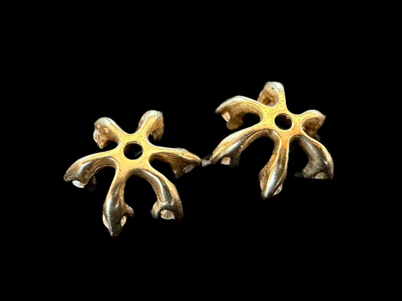 May include: Two gold-colored, star-shaped jewelry components with a central hole. The components have a unique, organic design with curved, petal-like extensions. The background is solid black, highlighting the metallic sheen of the objects.
