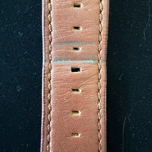 K&ouml;nnte beinhalten: Nahaufnahme eines braunen Leder-Uhrenarmbands mit gleichm&auml;&szlig;ig verteilten rechteckigen L&ouml;chern. Das Armband ist an den R&auml;ndern mit einem helleren braunen Faden gen&auml;ht. Das Leder weist Gebrauchsspuren auf.