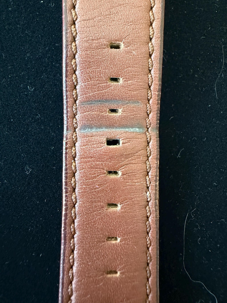 K&ouml;nnte beinhalten: Nahaufnahme eines braunen Leder-Uhrenarmbands mit gleichm&auml;&szlig;ig verteilten rechteckigen L&ouml;chern. Das Armband ist an den R&auml;ndern mit einem helleren braunen Faden gen&auml;ht. Das Leder weist Gebrauchsspuren auf.
