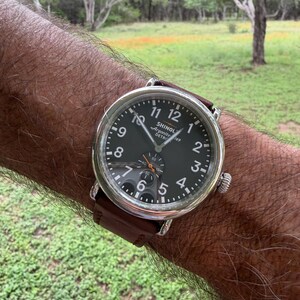 K&ouml;nnte beinhalten: Eine Armbanduhr mit dunklem Zifferblatt und silbernem Geh&auml;use wird gezeigt. Die Uhr hat ein braunes Lederarmband. Das Zifferblatt zeigt die Zahlen 1 bis 12, wobei der Markenname "SHINOLA" sichtbar ist. Die Uhr befindet sich am Handgelenk einer Person.