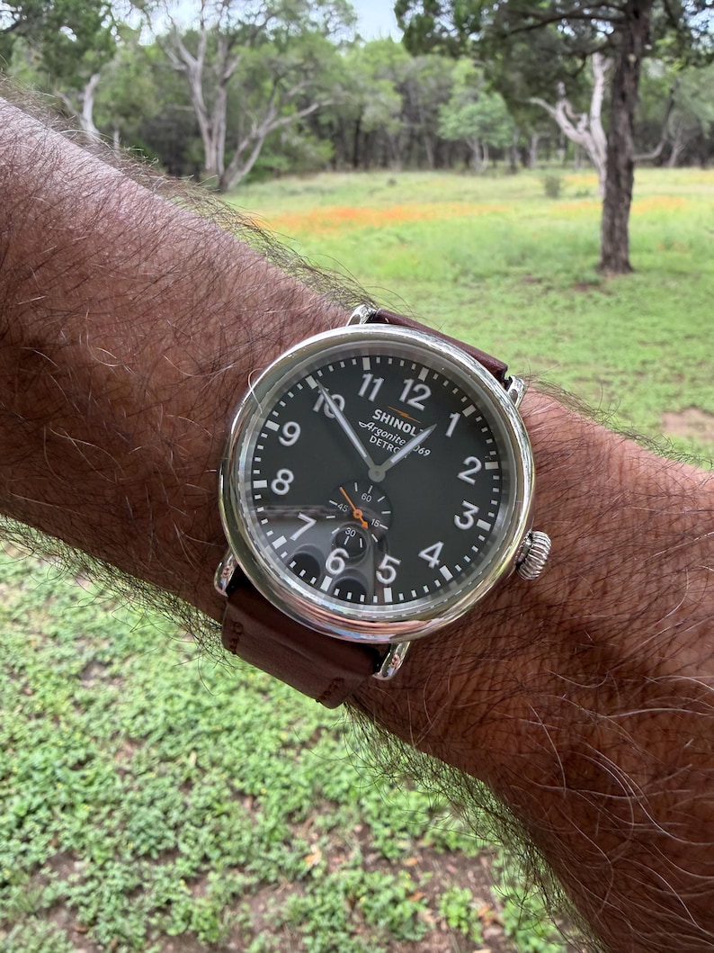 K&ouml;nnte beinhalten: Eine Armbanduhr mit dunklem Zifferblatt und silbernem Geh&auml;use wird gezeigt. Die Uhr hat ein braunes Lederarmband. Das Zifferblatt zeigt die Zahlen 1 bis 12, wobei der Markenname "SHINOLA" sichtbar ist. Die Uhr befindet sich am Handgelenk einer Person.