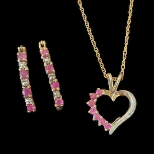 May include: A gold-toned jewelry set featuring a heart-shaped pendant necklace and matching hoop earrings. The pendant and earrings are adorned with pink gemstones and small clear stones, set against a black background.