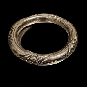 May include: A silver-colored ring with a textured, floral design. The ring is circular and appears to be made of metal. The design features a repeating pattern of leaves and flowers, creating a decorative band.