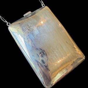 May include: A rectangular silver-toned metal case with a polished finish, featuring fine vertical line engravings and a circular monogram detail. The case is attached to a thin silver-toned metal chain and includes a small rectangular clasp closure.