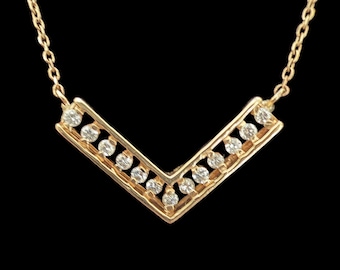 May include: A gold V-shaped pendant necklace featuring a series of small, round, clear stones set along the top edge of the chevron-shaped bar. The pendant is suspended from a delicate gold chain that attaches to both ends of the V-shaped metal frame.