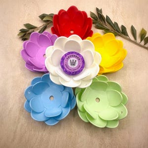 May include: A collection of colorful, flower-shaped soap dishes in red, purple, white, yellow, blue, and green. A lavender-scented soap is in the center of the white dish. The text on the soap reads "Lavender Sweet All Day Long."