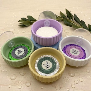 Op de afbeelding: Een verzameling kleine, kleurrijke keramische kommen met zuignappen. Elke kom bevat een verpakte zeep of badproduct. De labels luiden "Verbena", "Eucalyptus" en "Lavendel". De kommen zijn in groen, geel, paars en wit.