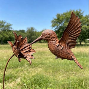 May include: A copper-coloured metal garden stake featuring a hummingbird in flight with its beak touching a multi-petaled flower. The sculpture has a textured, weathered finish and is mounted on a thin metal rod for placement in the ground.