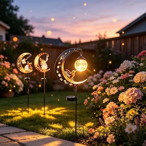 May include: Three solar garden stakes featuring crescent moon shapes with cut-out star patterns. Each stake holds a glowing, textured glass orb light. The foreground stake displays a hummingbird silhouette, while the others feature a fairy and a cat.
