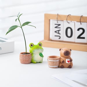 May include: A green frog figurine holding a small plant in a woven-style pot and a brown capybara figurine holding a small mug with a red heart design, positioned in front of a wooden desk calendar displaying the text JAN 02.
