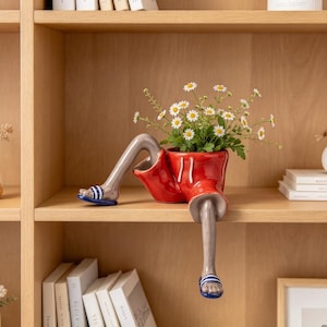 May include: A ceramic planter shaped like a pair of legs wearing red shorts and blue and white striped sandals. The planter is filled with a bouquet of white daisies with yellow centers, positioned to look as if the legs are dangling.