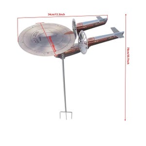 May include: A metal starship garden spinner featuring a circular saucer section with NCC-1701 markings, two cylindrical engine nacelles, and a vertical support stake. Dimensions are 34 cm wide, 18 cm deep, and 78 cm in total height.