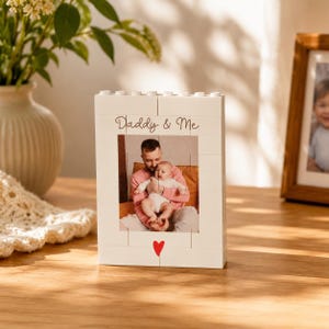 Puede incluir: Un marco de fotos blanco con el texto "Daddy & Me" y un corazón rojo. El marco contiene una foto de un padre y un bebé. Un jarrón con flores y una foto enmarcada están en el fondo.