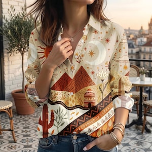 May include: Beige button-down shirt with a desert-themed print. The design includes a sun, moon, mountains, and geometric patterns in shades of brown, orange, and white. The shirt has long sleeves and a collared neckline.
