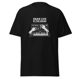 Può includere: Una t-shirt nera a maniche corte con una stampa grafica bianca di un sintetizzatore e mani che regolano i comandi. Sopra il sintetizzatore, il testo GRAB LIFE BY THE KNOBS è scritto in un carattere bianco in grassetto con linee orizzontali.
