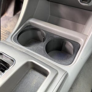 May include: A black, textured dual cup holder insert designed for a vehicle center console. The insert features two circular openings with vertical slots, positioned in front of a gray plastic console area with USB ports.