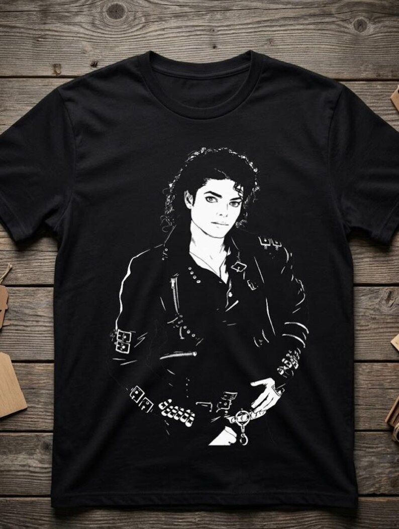 May include: A black short-sleeved t-shirt featuring a high-contrast white graphic print of a person with curly hair wearing a leather jacket adorned with multiple buckles, zippers, and metallic hardware details across the chest and sleeves.