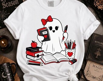 May include: A white short-sleeve t-shirt featuring a graphic of a cartoon ghost with a red bow, holding a notebook and pen. The ghost is surrounded by a stack of books, an apple, a pencil holder with pencils, and an open book in red and black.
