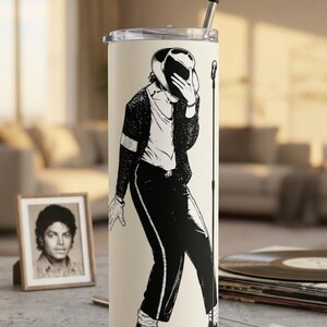 May include: A tall, cylindrical, cream-colored tumbler featuring a black graphic illustration of a performer in a fedora, sequined jacket, and trousers, posing with a microphone stand. The tumbler has a clear lid and a metal straw inserted.
