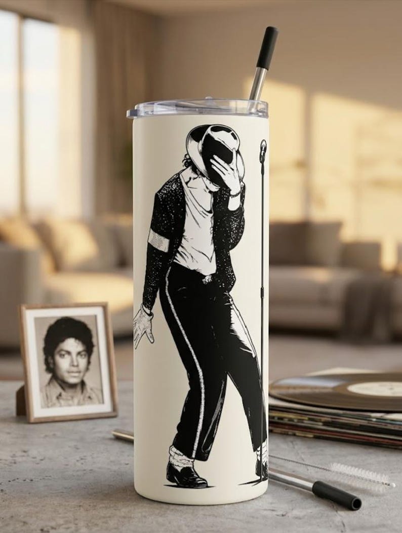 May include: A tall, cylindrical, cream-colored tumbler featuring a black graphic illustration of a performer in a fedora, sequined jacket, and trousers, posing with a microphone stand. The tumbler has a clear lid and a metal straw inserted.