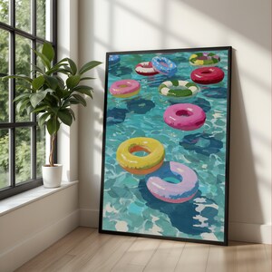 May include: A framed wall art print depicting a swimming pool filled with multiple colourful inflatable pool floats. The painting features various circular rings in shades of pink, yellow, blue, green, and red, floating on turquoise water.