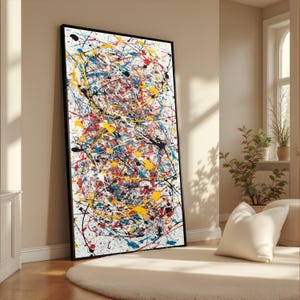 Op de afbeelding: Abstract expressionistisch schilderij met een zwarte lijst. Het kunstwerk toont een witte achtergrond met kleurrijke verfspetters in geel, rood, blauw en zwart. Het schilderij leunt tegen een beige muur in een lichte kamer.