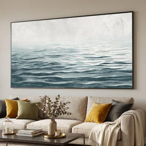 May include: A large rectangular framed artwork depicting a serene ocean scene. The artwork features shades of blue and white, with textured waves and a light, cloudy sky. The artwork is displayed above a beige sofa with colorful throw pillows.