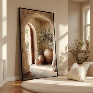 May include: A large, rectangular mirror with a black frame leans against a wall. The mirror reflects an arched doorway leading to a sunlit courtyard with terracotta pots and greenery. A white rug and pillow are in the foreground.