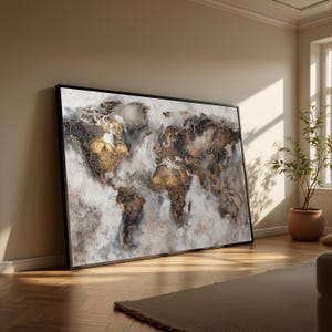 Op de afbeelding: Een ingelijste wereldkaart kunstafdruk. De kaart toont gouden en zwarte continenten tegen een witte en grijze achtergrond. Het kunstwerk wordt getoond in een kamer met natuurlijk licht en een houten vloer.