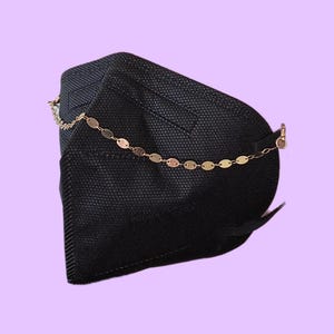 May include: A black face mask with a gold chain attached. The chain is made of small, flat, oval-shaped links. The mask is made of a textured fabric and has a curved shape.