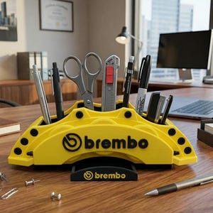 Może przedstawiać: Żółty organizer na biurko w kształcie zacisku hamulcowego z logo "brembo". Zawiera długopisy, nożyczki i nóż do cięcia. Organizer stoi na drewnianym biurku z innymi artykułami biurowymi.
