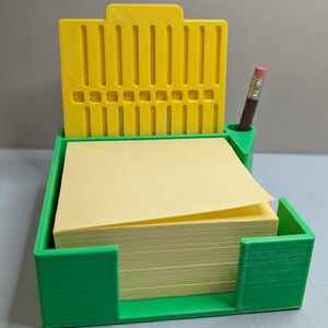 May include: A 3D-printed desk organiser in green and yellow. It holds a stack of yellow sticky notes and has a slot for a pencil. The back of the organiser is yellow with vertical slots.