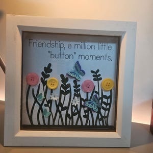 Puede incluir: Una obra de arte enmarcada con el texto "Friendship, a million little 'button' moments." Presenta un diseño floral con botones y mariposas. El marco es blanco y la obra tiene un fondo claro.