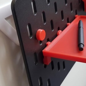 May include: A black pegboard with a red shelf and accessories. The shelf holds a black pen and silver paper clips. Red pegs secure the shelf. The pegboard is mounted on a white support.