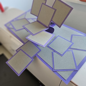 Könnte beinhalten: Ein handgemachtes Papierbastelprojekt mit grauen quadratischen Karten mit lila Rändern, von denen einige angehoben sind, wodurch ein 3D-Effekt entsteht. Die Karten sind auf einer größeren Basis mit lila Rand angeordnet, was auf ein Pop-up-Design hindeutet.