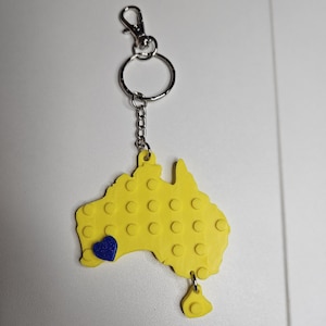 Puede incluir: Un llavero amarillo con la forma de Australia, con un corazón azul y una pequeña pieza amarilla colgando. Tiene un mosquetón y un anillo plateados.